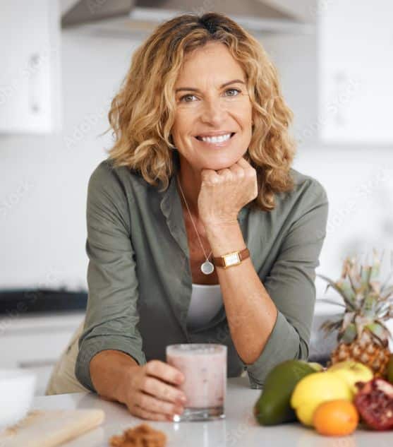 A woman with wavy blonde hair standing at her kitchen counter drinking coffee and smiling