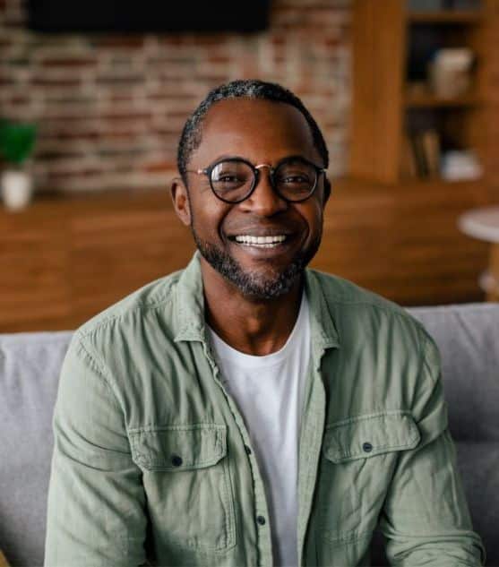A mature man in glasses and a green button-up sitting on a couch and smiling